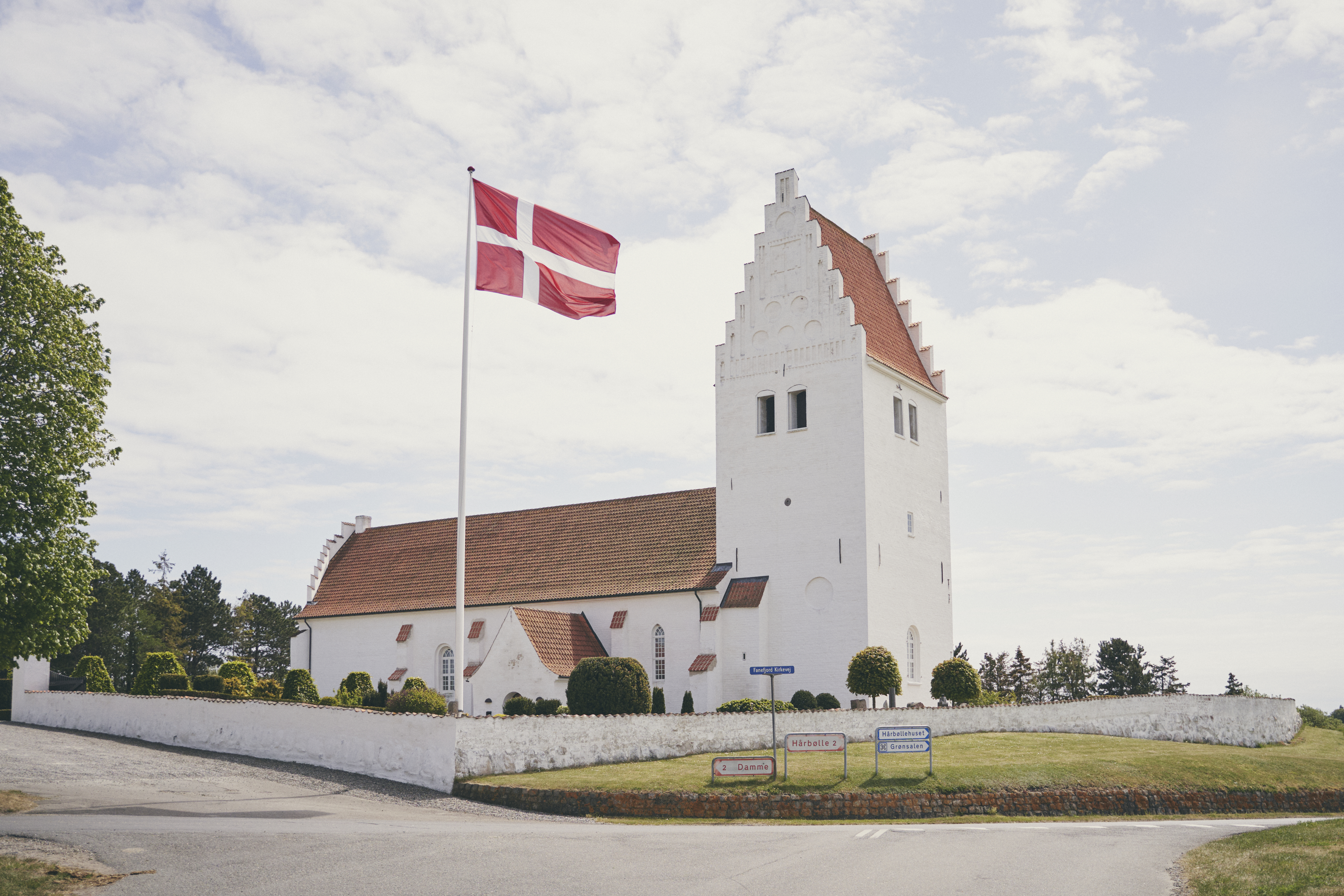 Fanefjord Kirke set fra vejen, med lyseblå himmel og hejst dannebro der vejrer i vinden